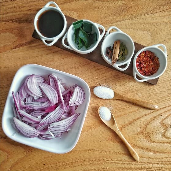 Ingredients on a table: Sliced onion,Chili flakes,cinnamon,cloves,cardamom,pandan leaf,curry leaves, Brindle berry paste.